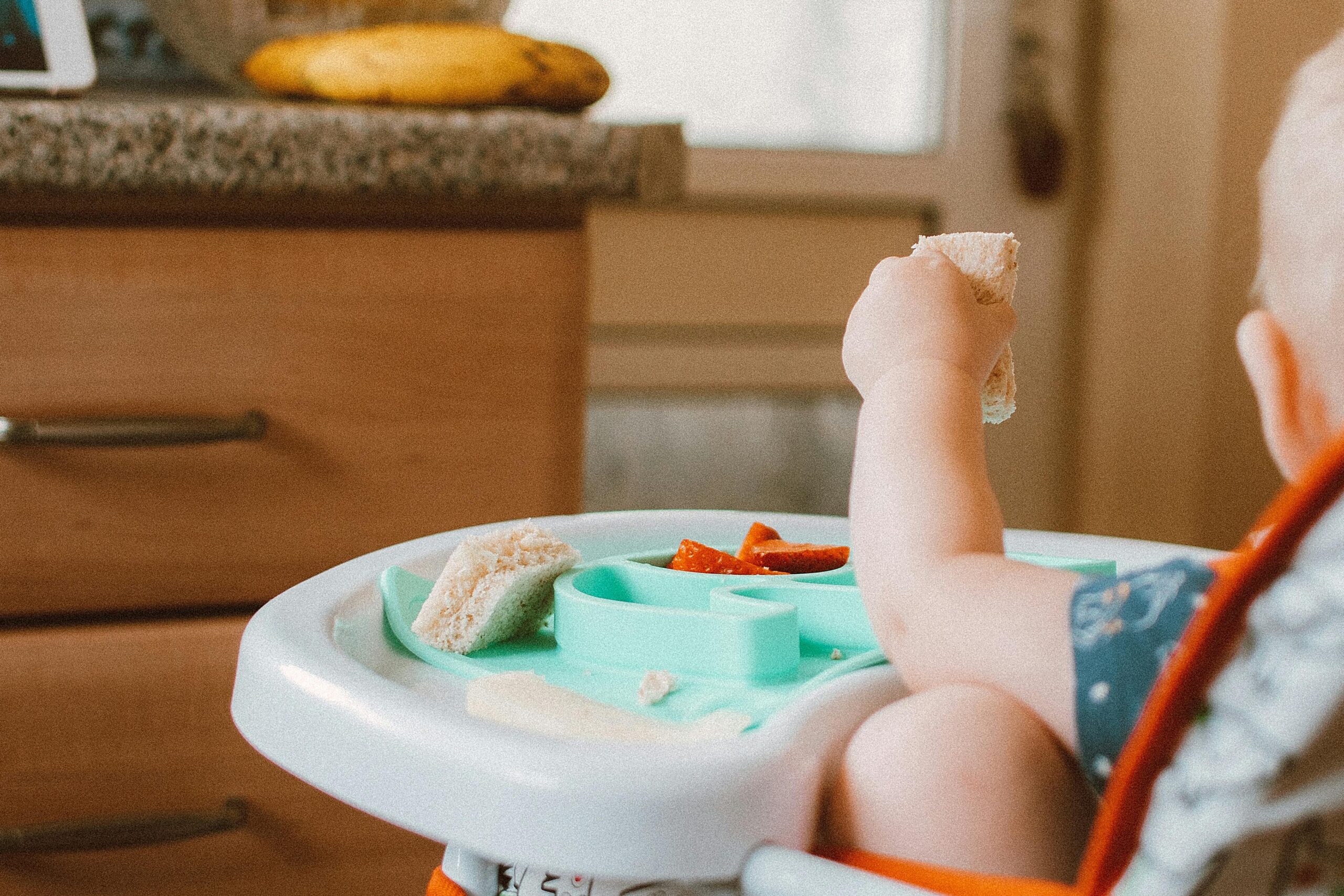 Baby in high chair eating food, surrounded by bananas in a kitchen setting.