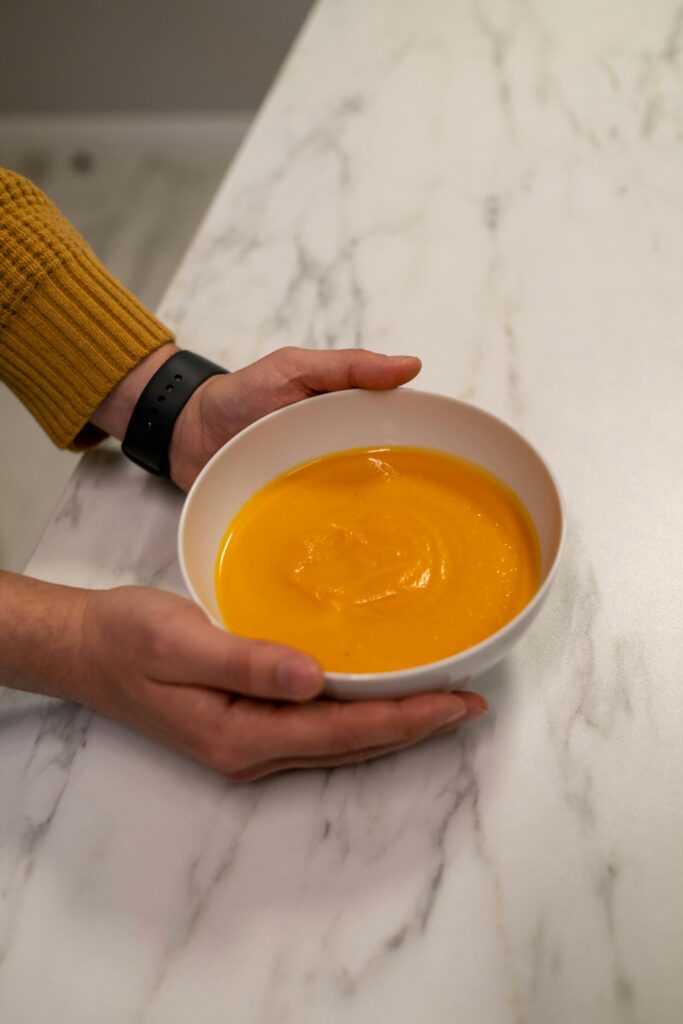 Warm pumpkin soup served in a ceramic bowl held by hands over a marble surface.