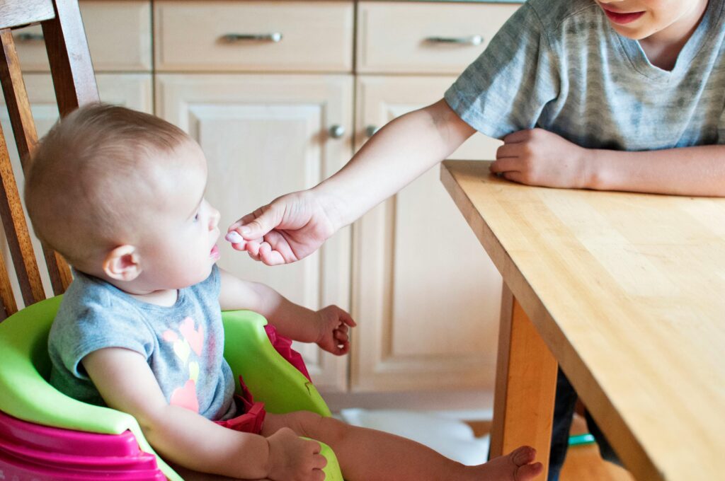 A cute baby in a high chair being fed by an older child in a bright kitchen setting.